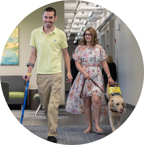 Two coworkers walking down the hall together. The man has a cane and the woman has a guide dog.