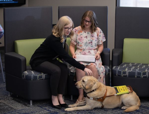 Two young businesswomen sitting in chairs. One woman is reading notes and the other is petting her guide dog.
